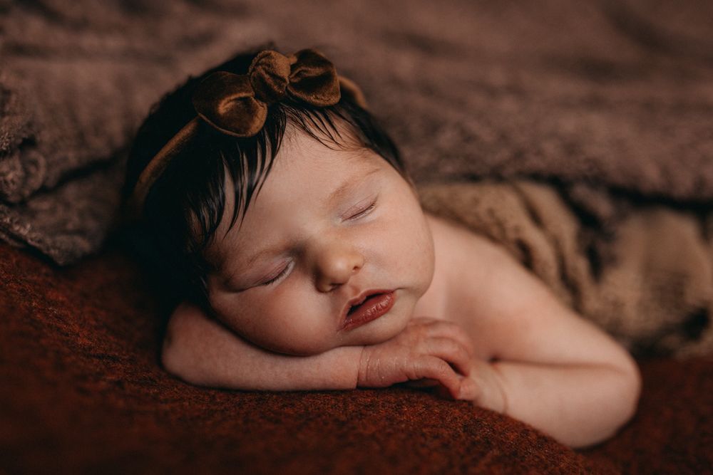 Sleeping newborn baby girl with dark hair wearing a brown bow headband, wrapped in a soft brown blanket.