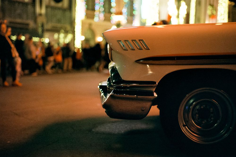 Close-up view of a vintage car's front end at night with blurred lights and people in the background.