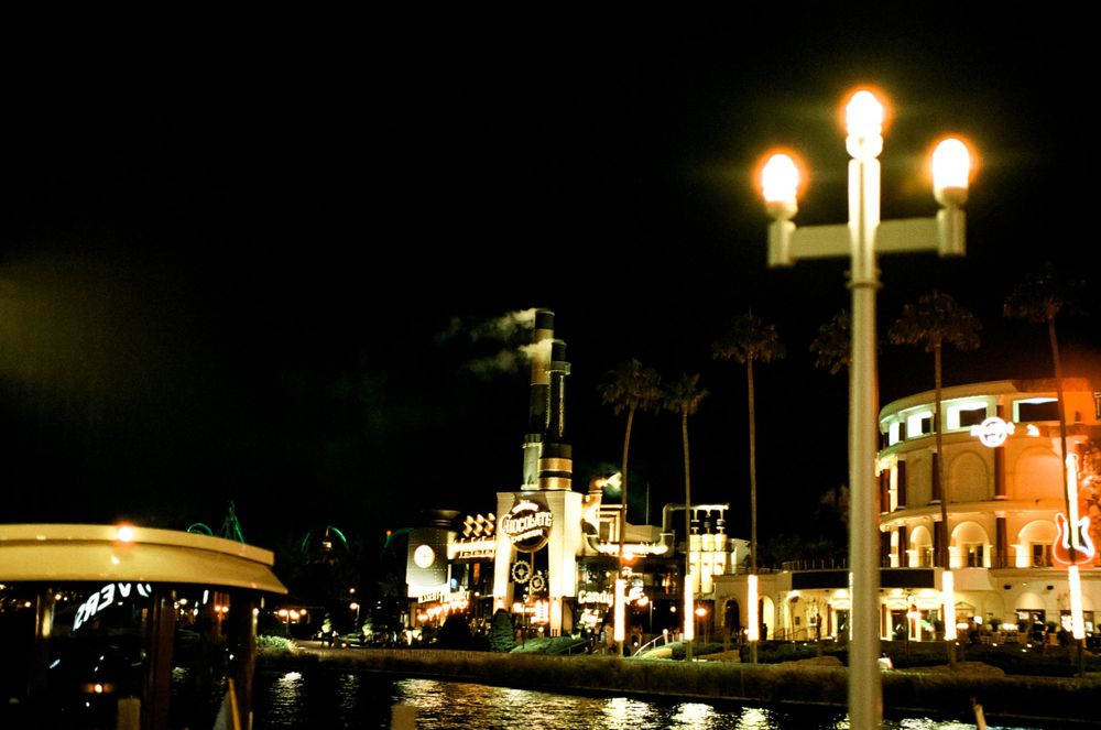 Nighttime view of a waterfront with illuminated buildings and ornate lamp posts.
