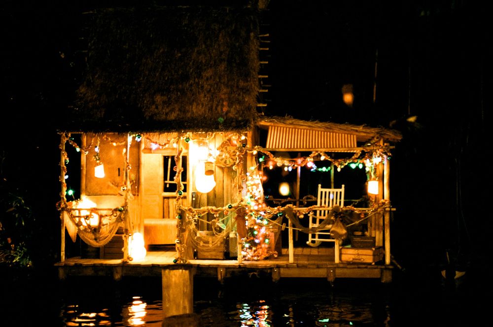 A cozy houseboat decorated with warm glowing Christmas lights reflects on calm water at night.