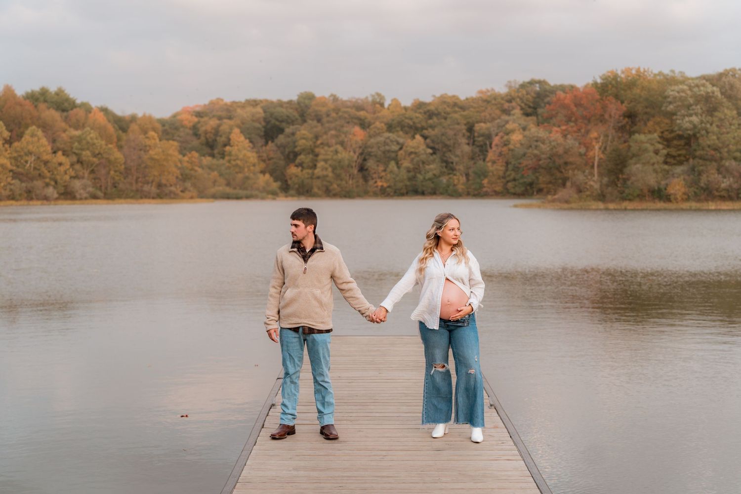 Two people standing on a wooden dock holding hands by a calm lake during autumn with colorful fall foliage in background.
