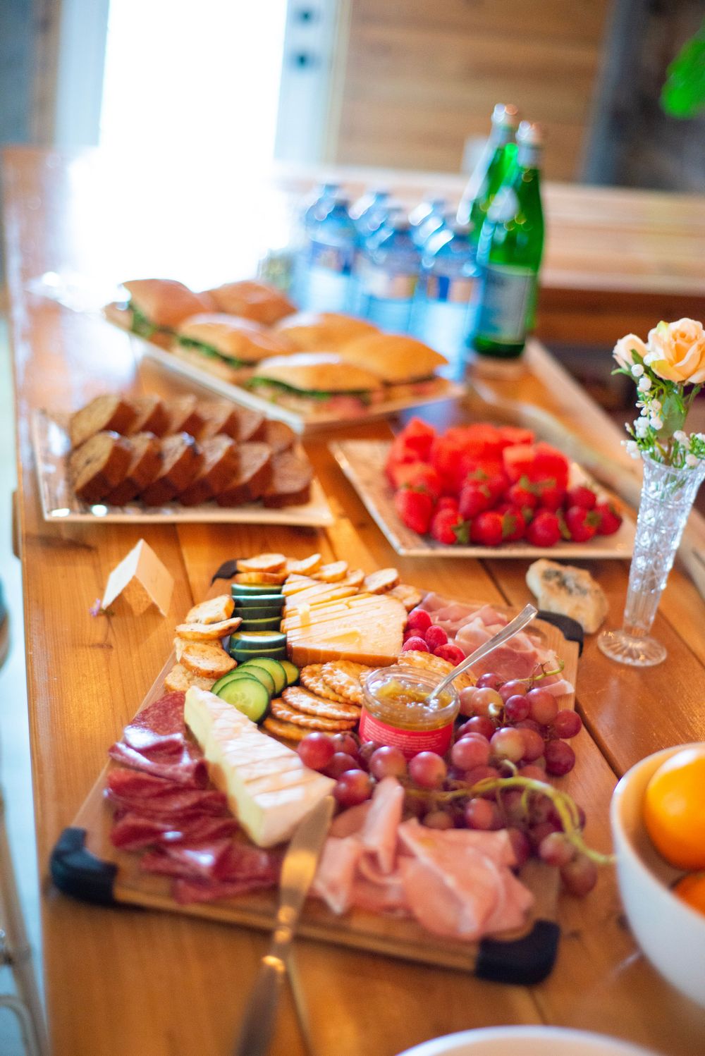 A charcuterie board displays meats, cheeses, grapes, and sandwiches alongside beverages on a wooden table.