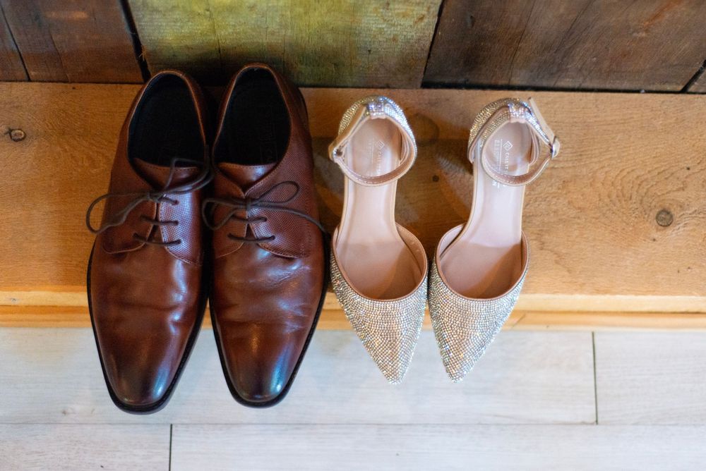 A pair of brown leather dress shoes next to glittery beige women's heels arranged on wooden floorboards.
