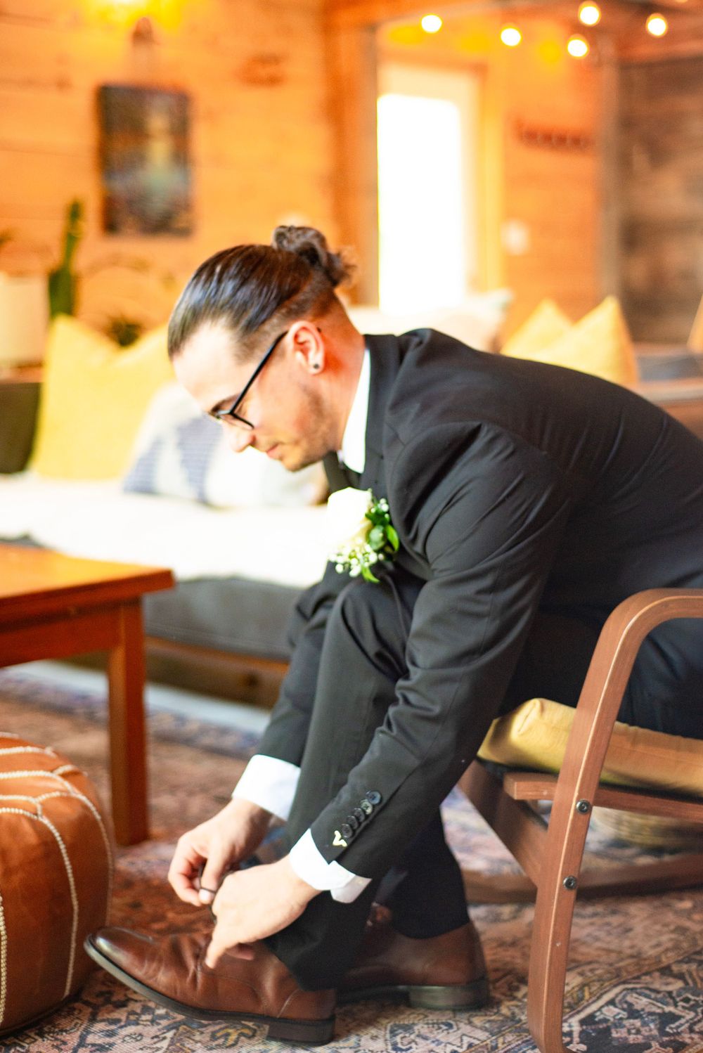 Groom in black suit and glasses adjusts his brown shoes while getting ready in a warmly lit rustic room.