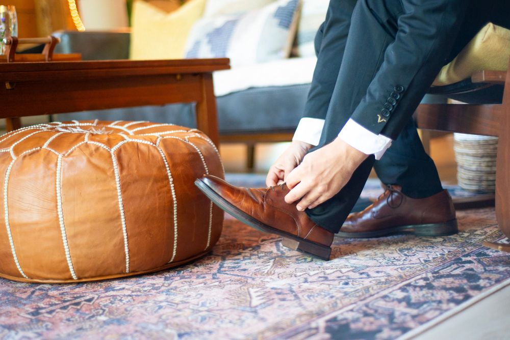 Person in formal attire putting on brown leather dress shoes next to a leather ottoman on patterned rug.