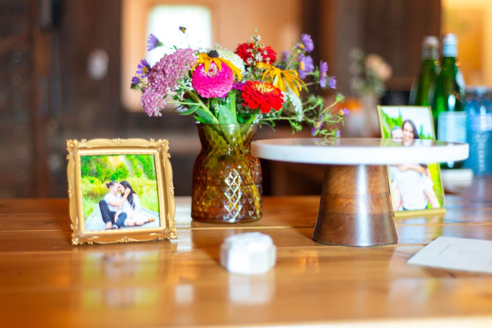 A bright floral arrangement in a glass vase sits on a wooden table next to a framed photograph and table decorations.