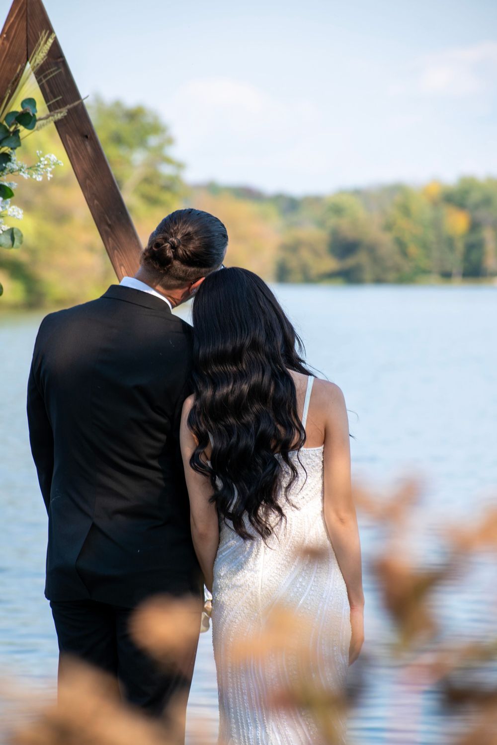 A couple in wedding attire stands together overlooking a peaceful lake on an autumn day.