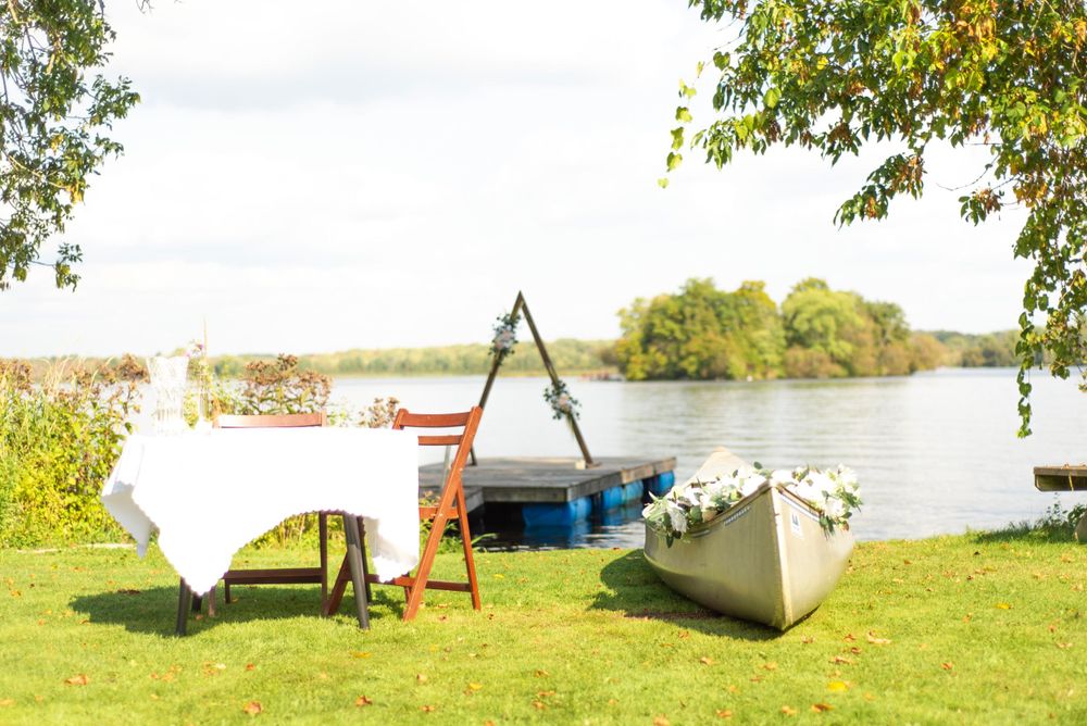 A rustic outdoor wedding setup with white tables and chairs arranged on grass near a waterfront.
