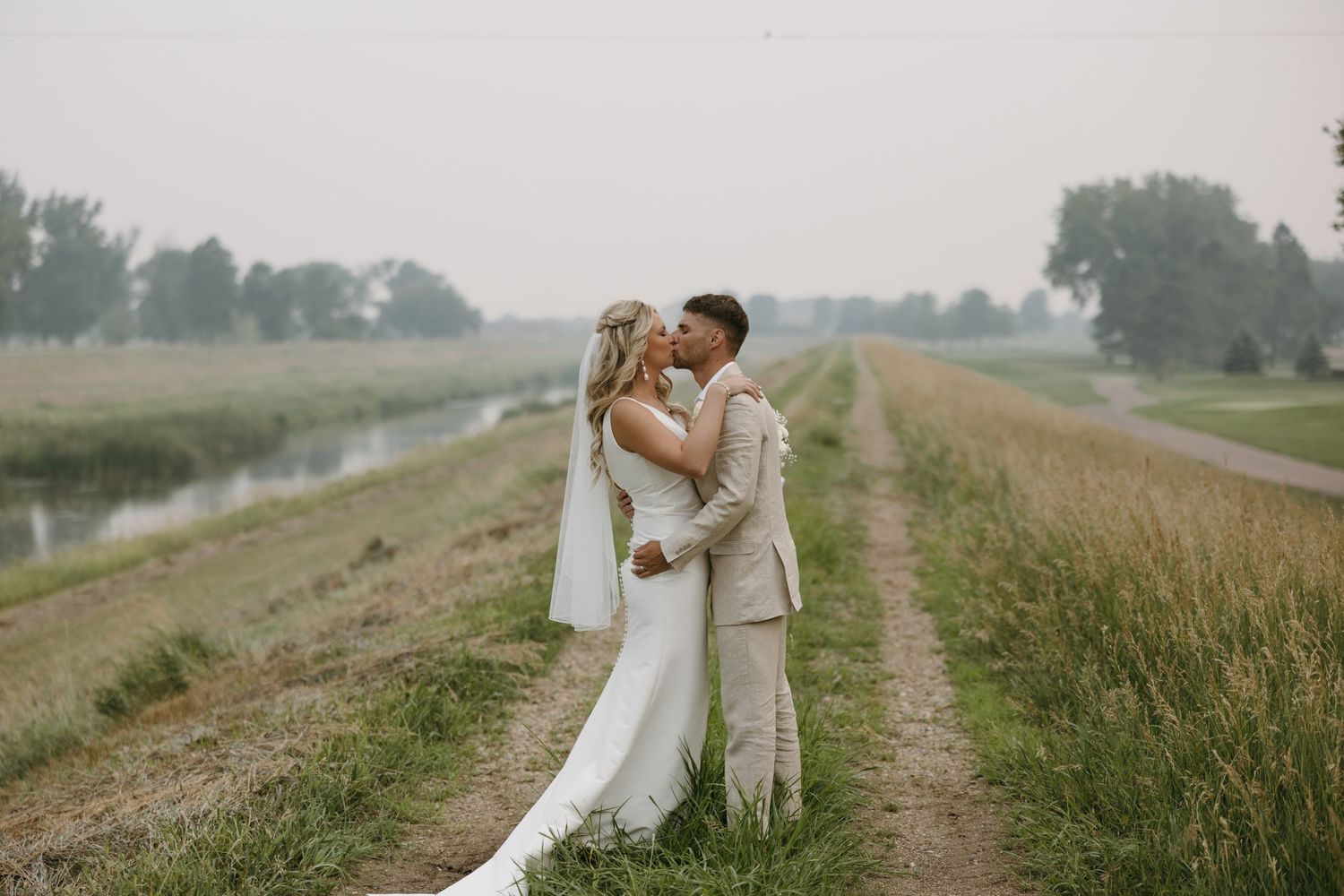 Couple in wedding attire share intimate moment on rural path next to canal at sunset.