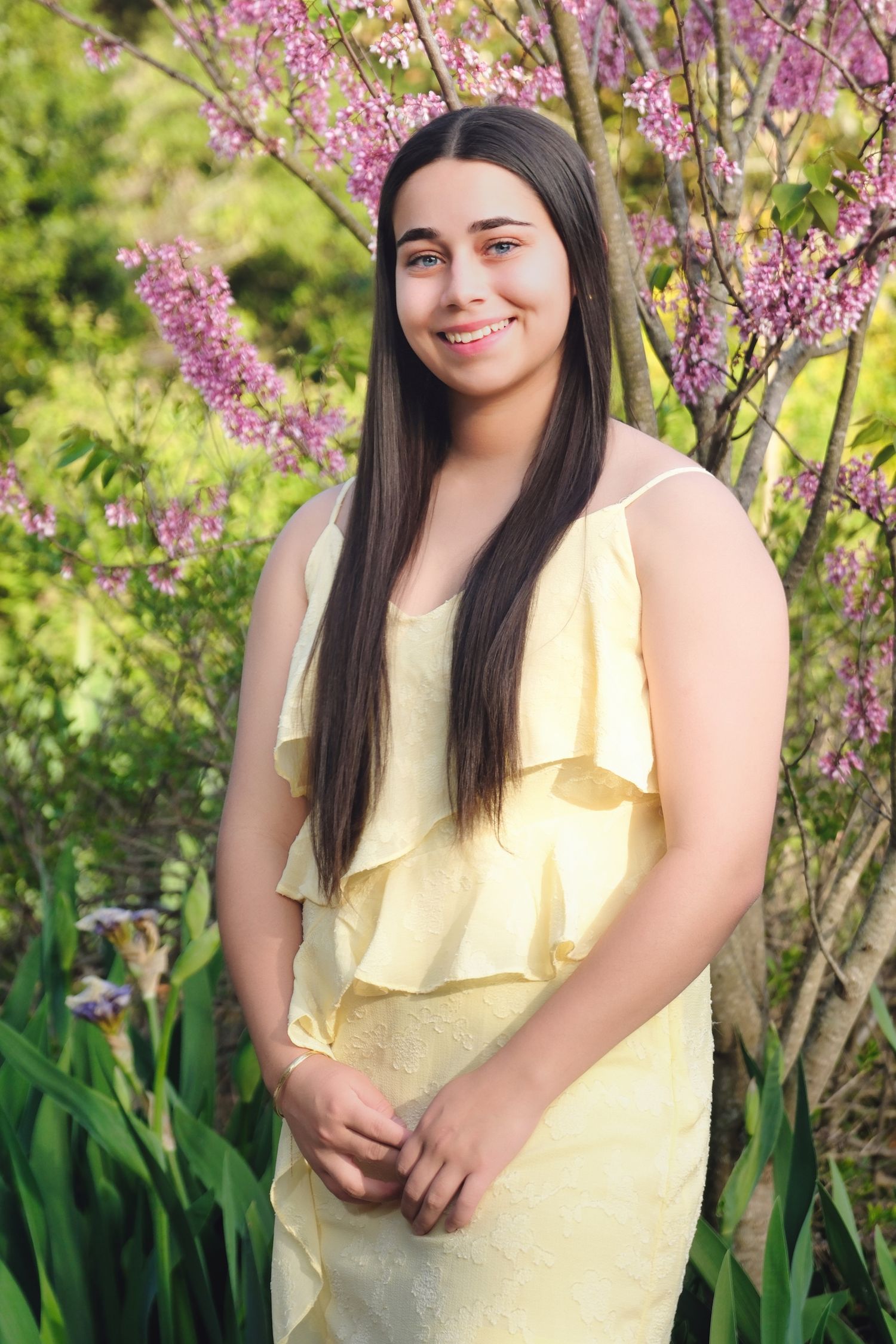 A figure in a pale yellow ruffled dress poses outdoors against blooming pink spring flowers and green foliage.