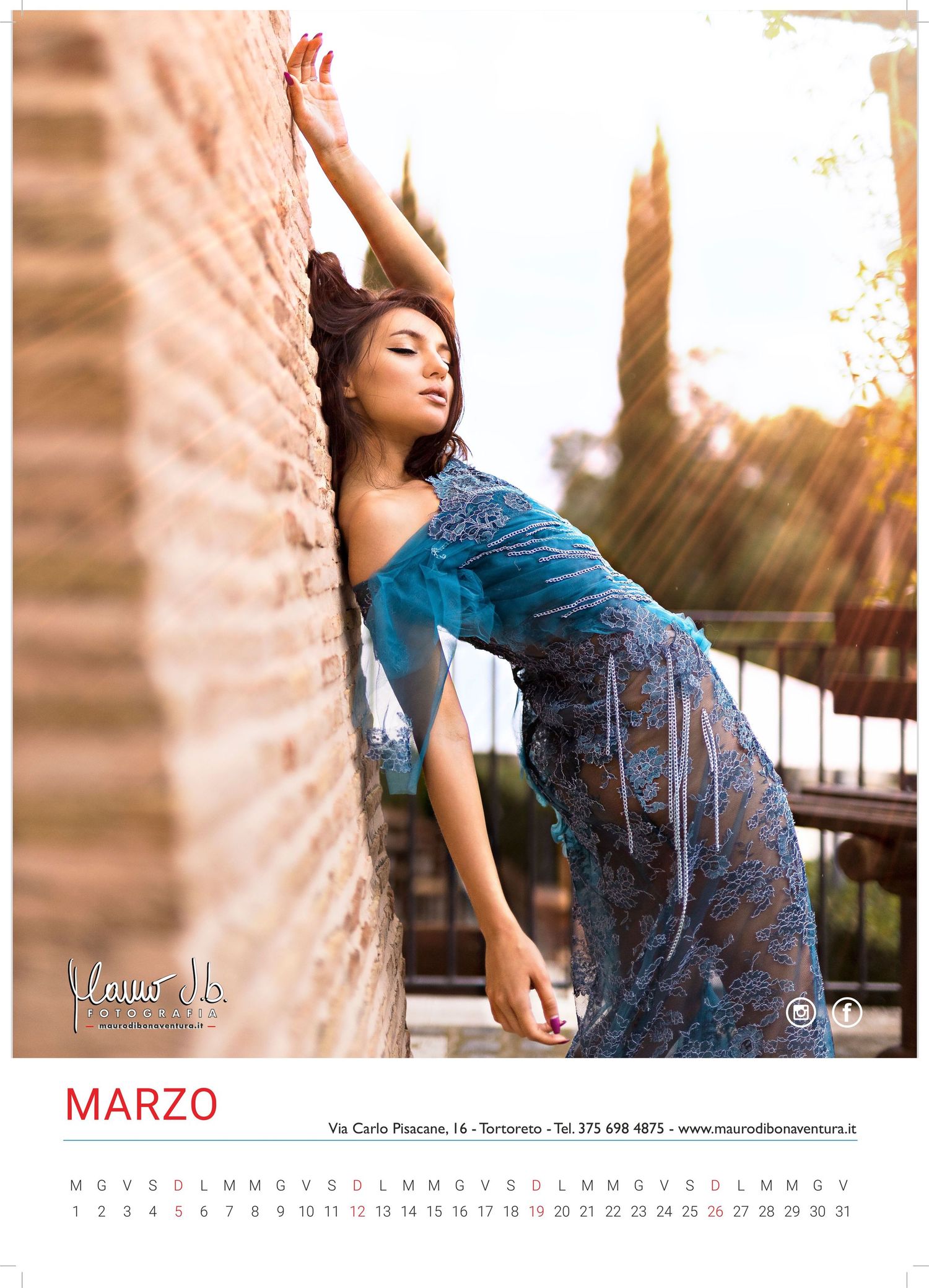 Fashion model in blue dress poses dramatically against brick wall during sunset photo shoot for March calendar.