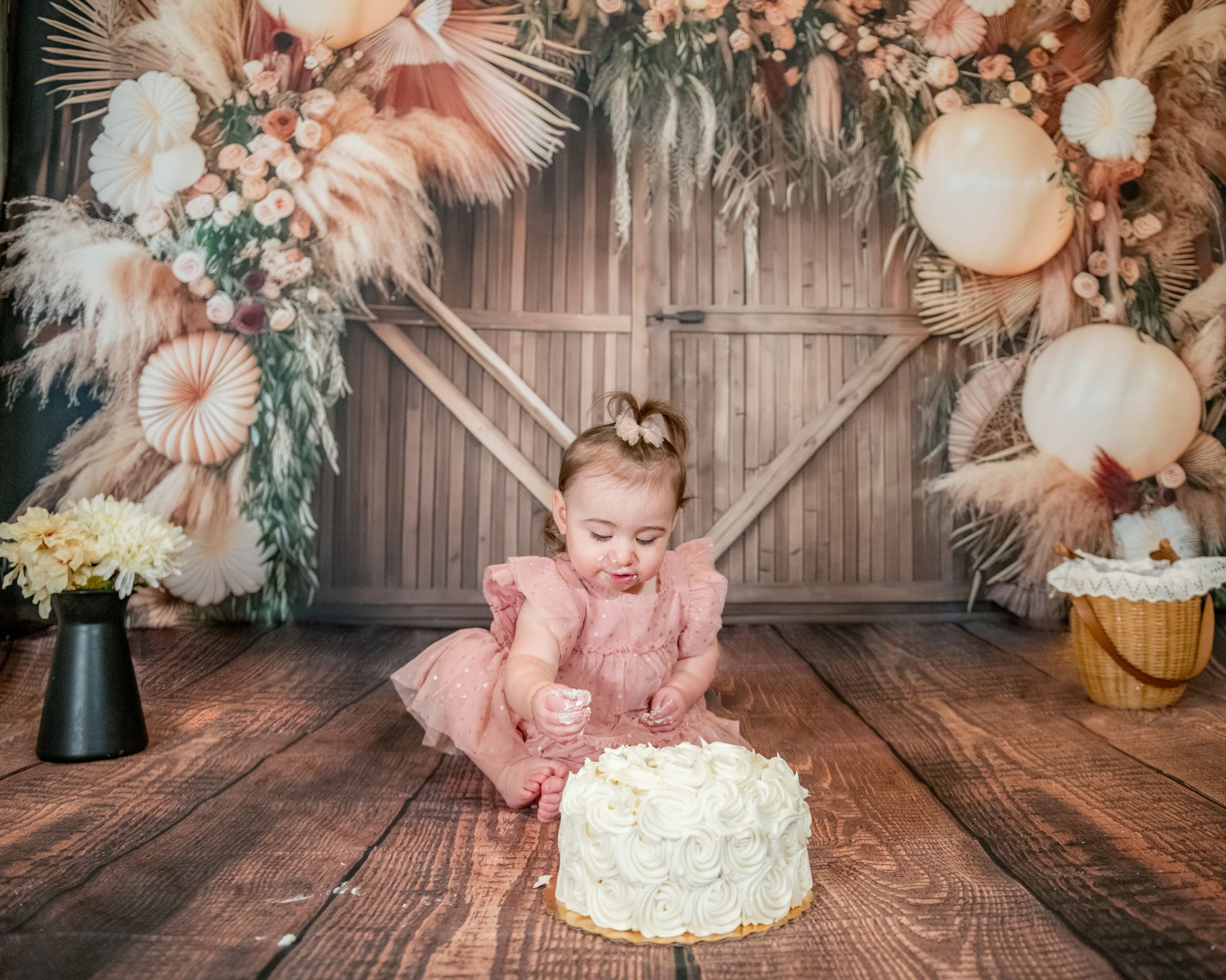 Rustic cake smash photo setup with pampas grass, balloon garland and white cake against wooden backdrop in peach tones.