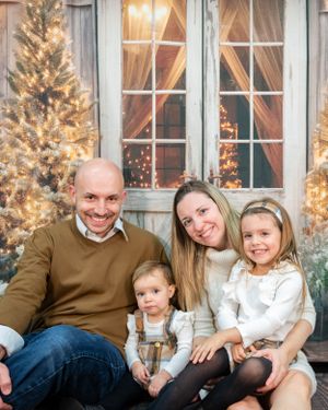 A family sits together outdoors for a cozy holiday portrait with lit Christmas trees and windows glowing behind them.