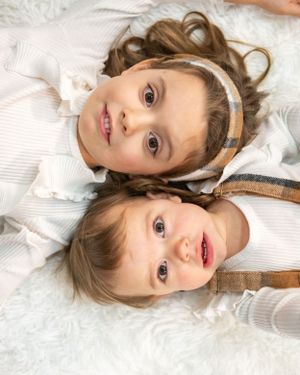 Two children lying together on a white background wearing light-colored clothing and looking up at the camera.