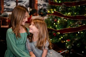 Two girls in green and gray dresses share a sweet moment in front of a decorated Christmas tree.