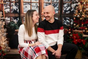 A couple in festive clothing sits together near a decorated Christmas tree by a window.