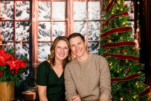 A couple sits together by a Christmas window display with a decorated tree and poinsettias in a cozy holiday setting.