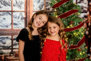 Two children wearing festive holiday outfits pose in front of a decorated Christmas tree and snowy window backdrop.