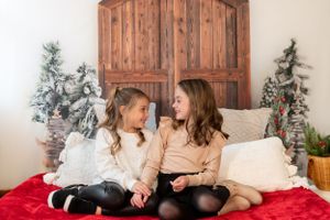 Two children sit together on a couch with holiday decor and snow-flocked trees in the background.