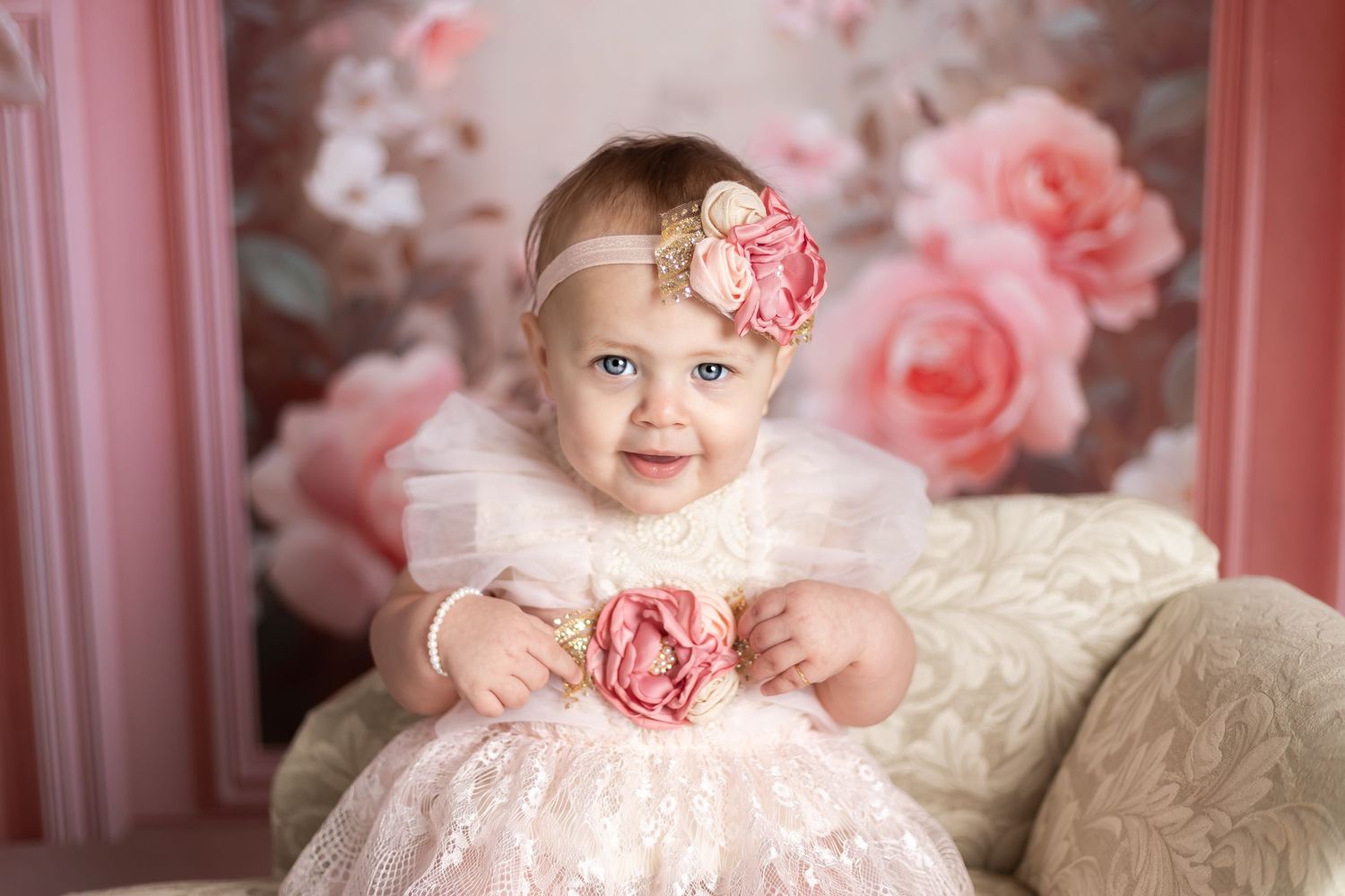 Baby in pink tulle dress with floral headband sits against pink rose backdrop for portrait session.