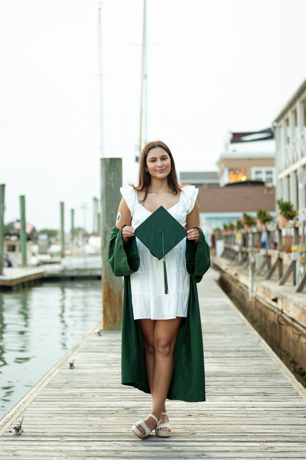 Graduate in white dress and green graduation gown poses with diploma on wooden dock by marina.