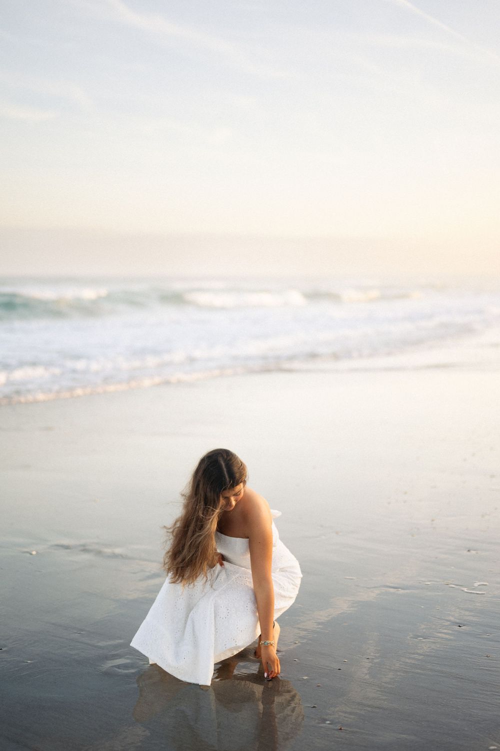 A figure in a white dress sits contemplatively on a misty beach shoreline at sunset.