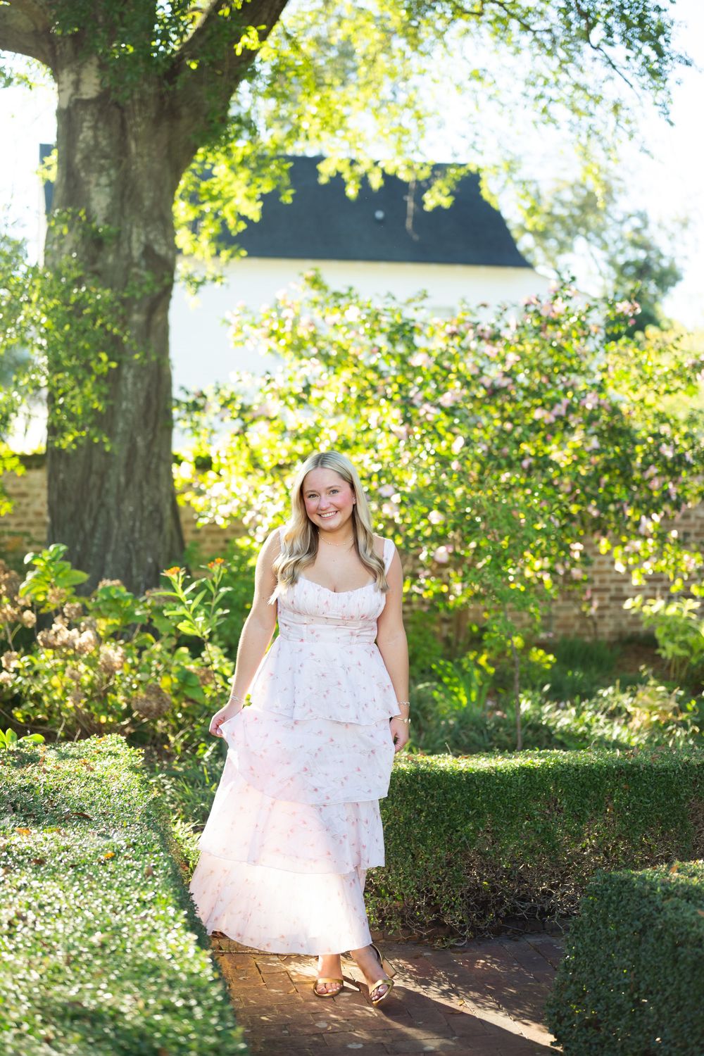 A person in a white lace dress poses outdoors in a sunlit garden setting with green trees and bushes in the background.