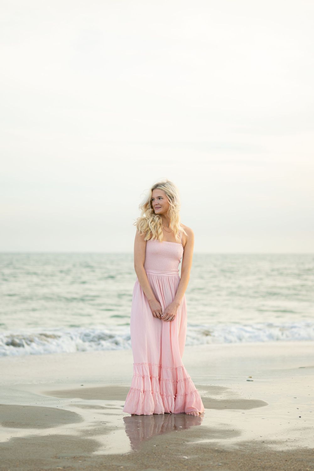 Figure in flowy pink maxi dress stands gracefully on beach shore at sunset with waves and ocean in background.