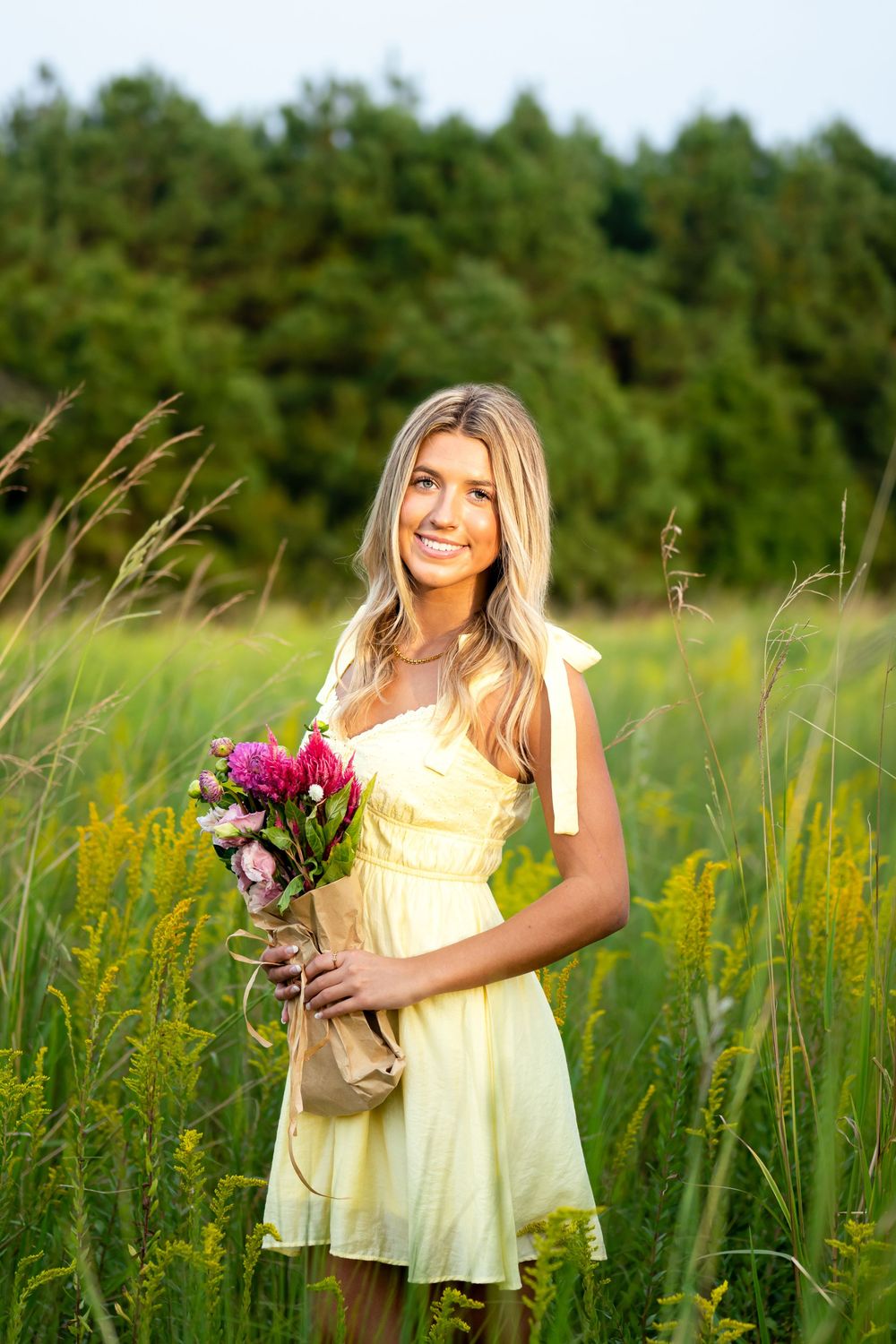 Someone in a yellow sundress holds a bouquet of pink flowers while standing in a sunlit meadow with tall grass.