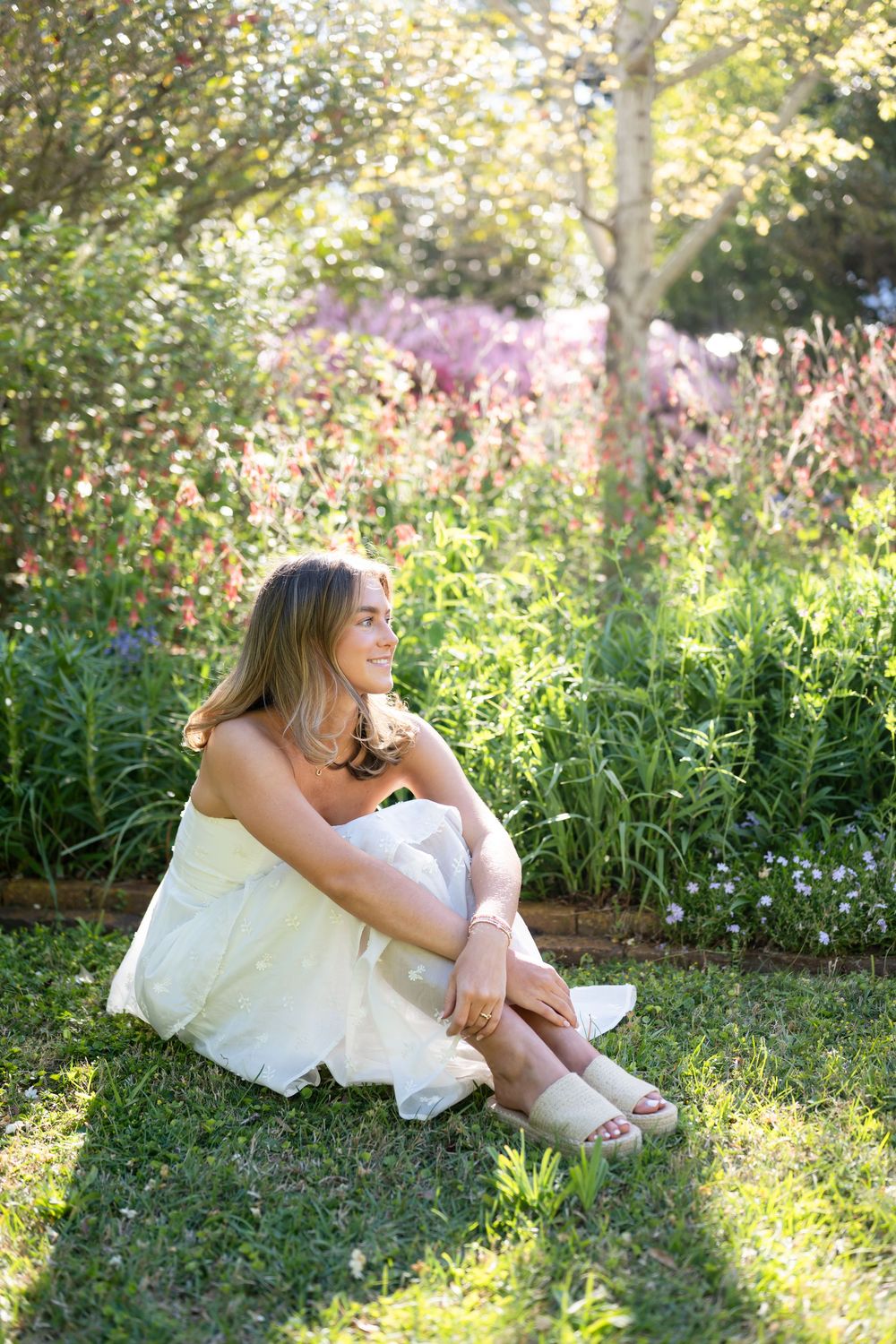 Young woman in white dress sitting on grass in a sunlit garden with colorful wildflowers blooming behind her.