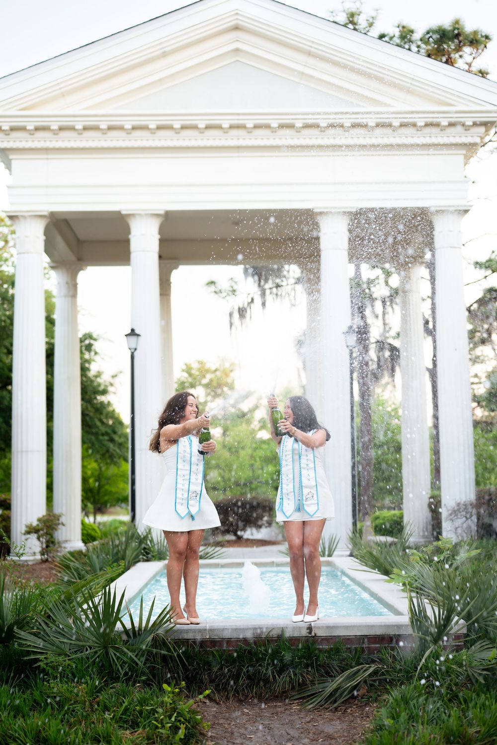 Two women in white dresses celebrate near a fountain by a white columned gazebo surrounded by lush greenery.