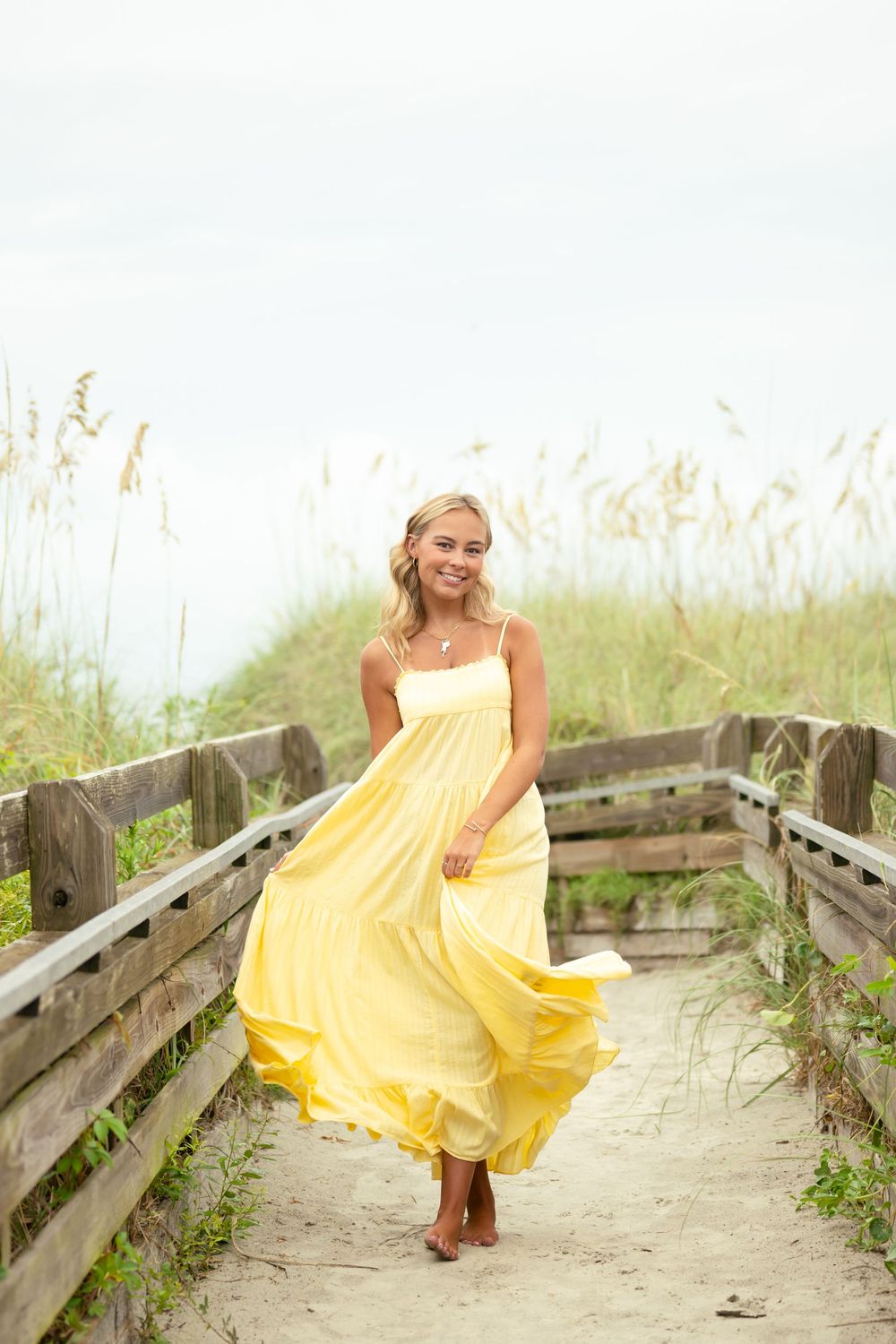 A yellow sundress flows in the breeze as someone poses on a wooden boardwalk among beach grass and sand dunes.