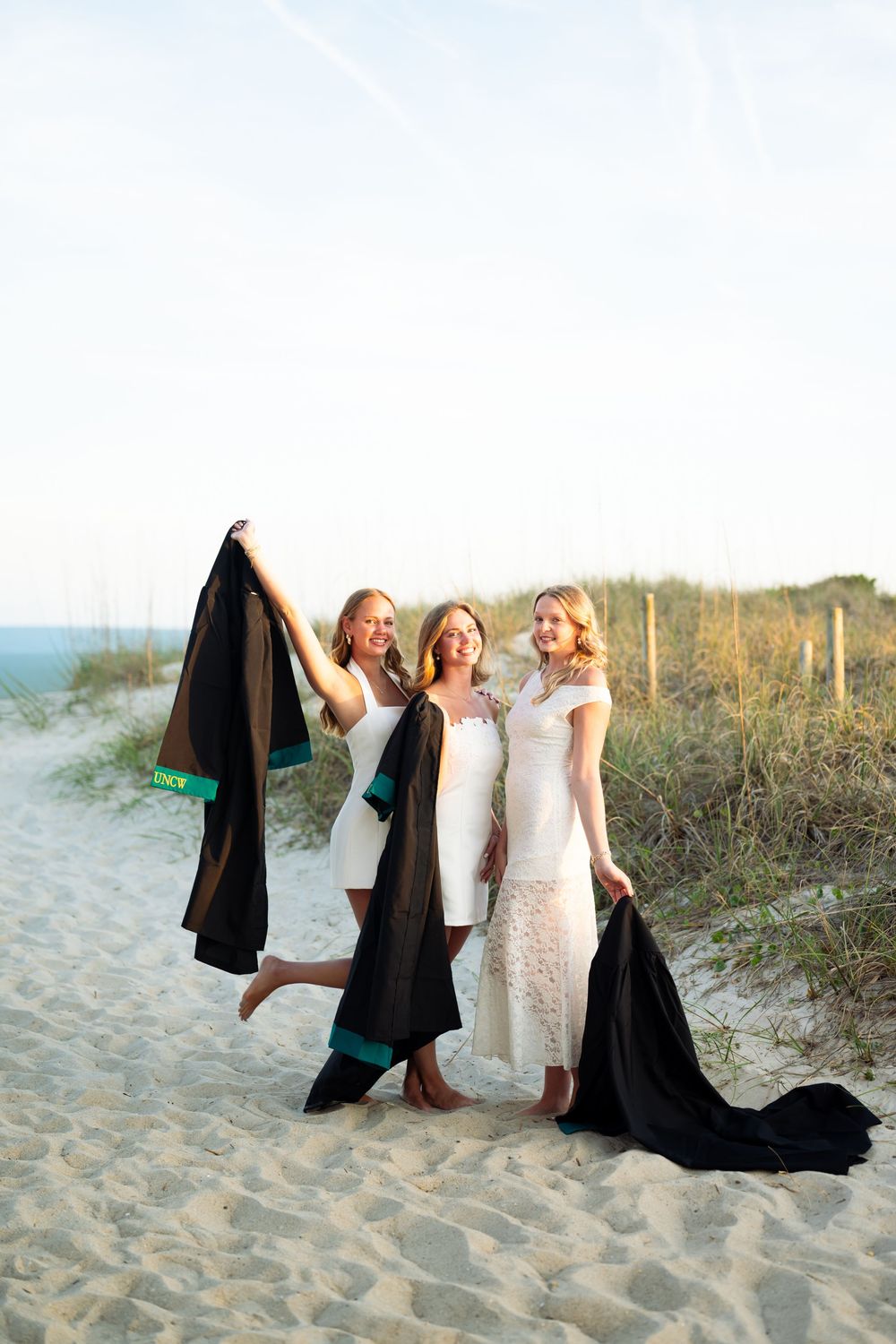 Three women in white dresses pose on a sandy beach holding black fabric, with dunes and fence in background.
