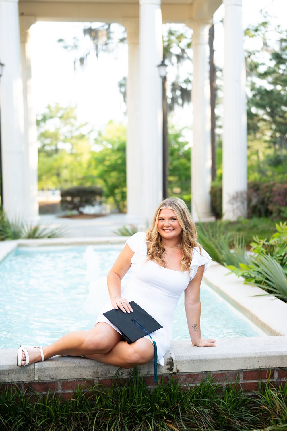 Smiling blonde graduate in white dress holding diploma by elegant pool with white columns outdoors.