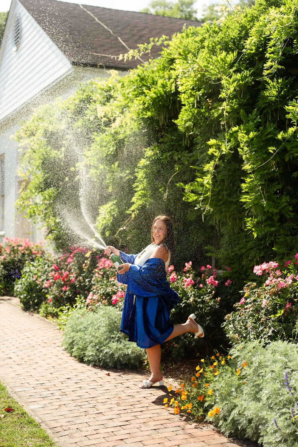 Woman in blue dress joyfully spraying water hose in a lush garden with roses and green hedges on a sunny day.