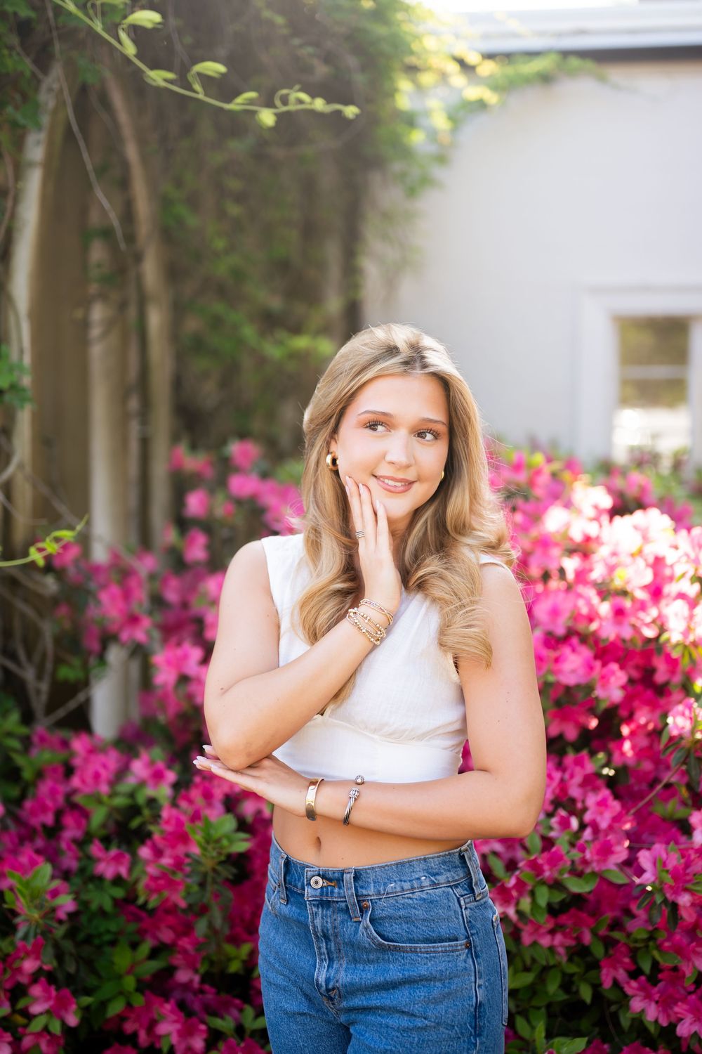 Young blonde woman in white crop top and jeans posing in front of vibrant pink azalea bushes outdoors.