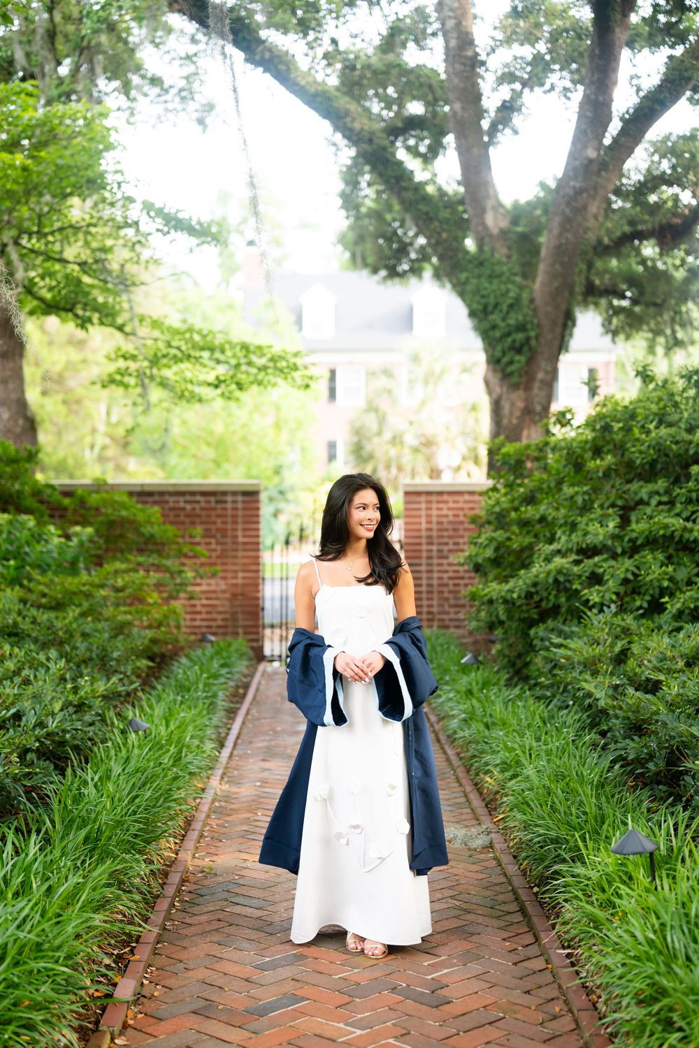 Woman in white gown and navy wrap poses on brick garden path surrounded by lush greenery and brick walls.
