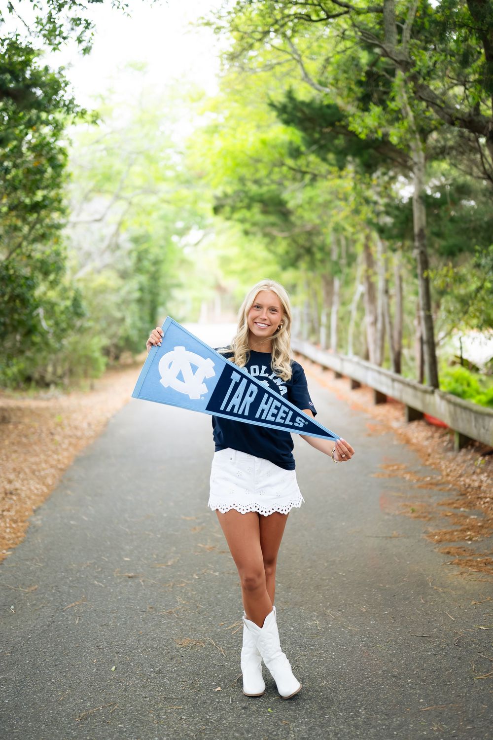 Smiling blonde woman in UNC Tar Heels shirt holding pennant flags on a tree-lined path.