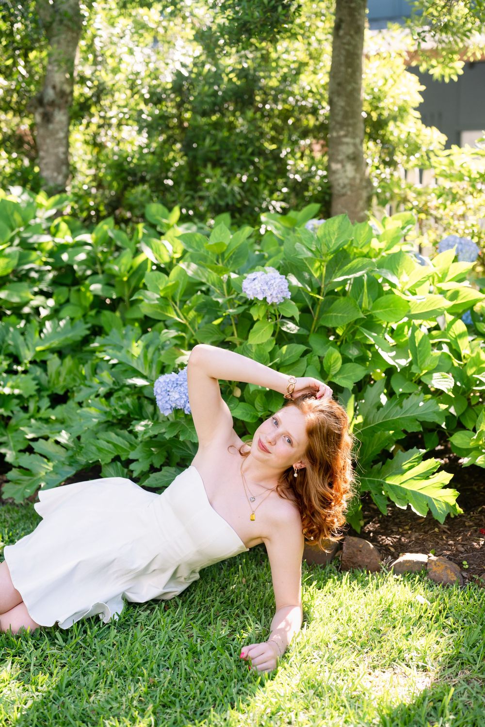 Red-haired woman in white dress lying on green grass beside blooming hydrangeas in a sunny garden.