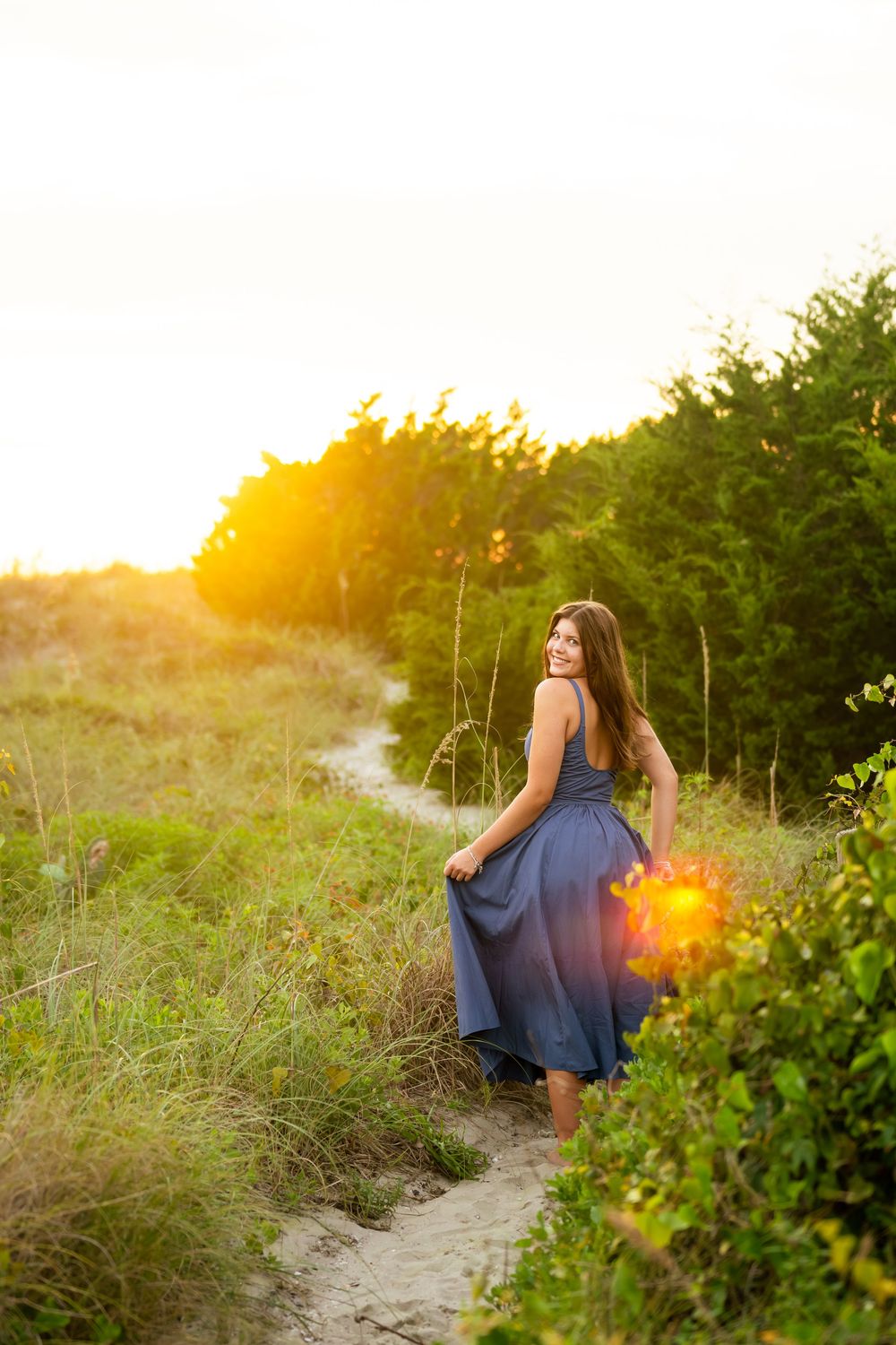 A person in a flowing blue dress walks along a sandy beach path at sunset with sunlit foliage in the foreground.
