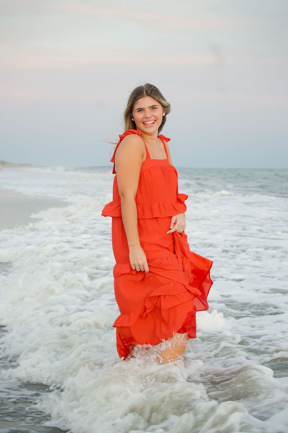 Person in a flowing red dress stands in ocean waves at the beach during sunset.