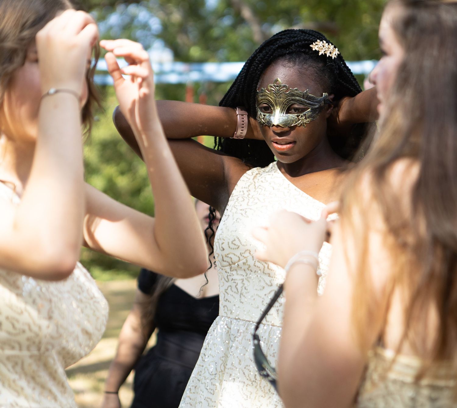 People help adjust a sparkly masquerade mask during an outdoor celebration.