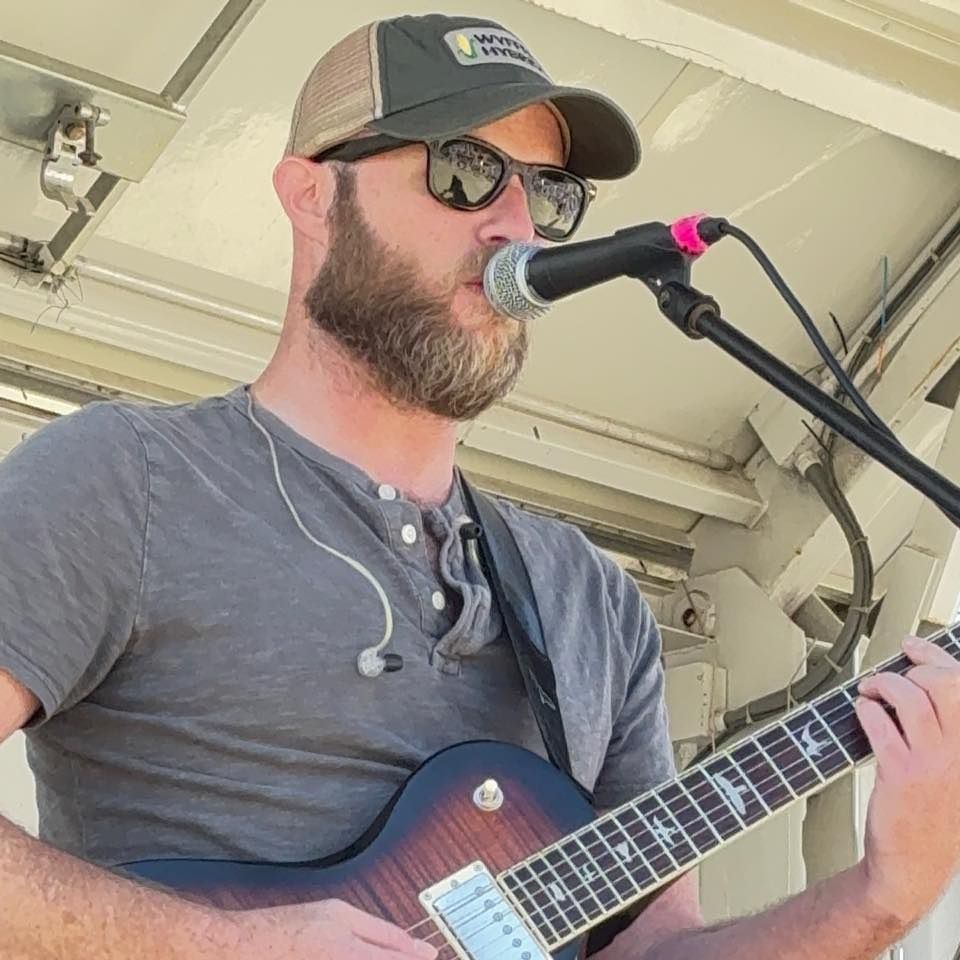 Musician performing with guitar and microphone wearing baseball cap.
