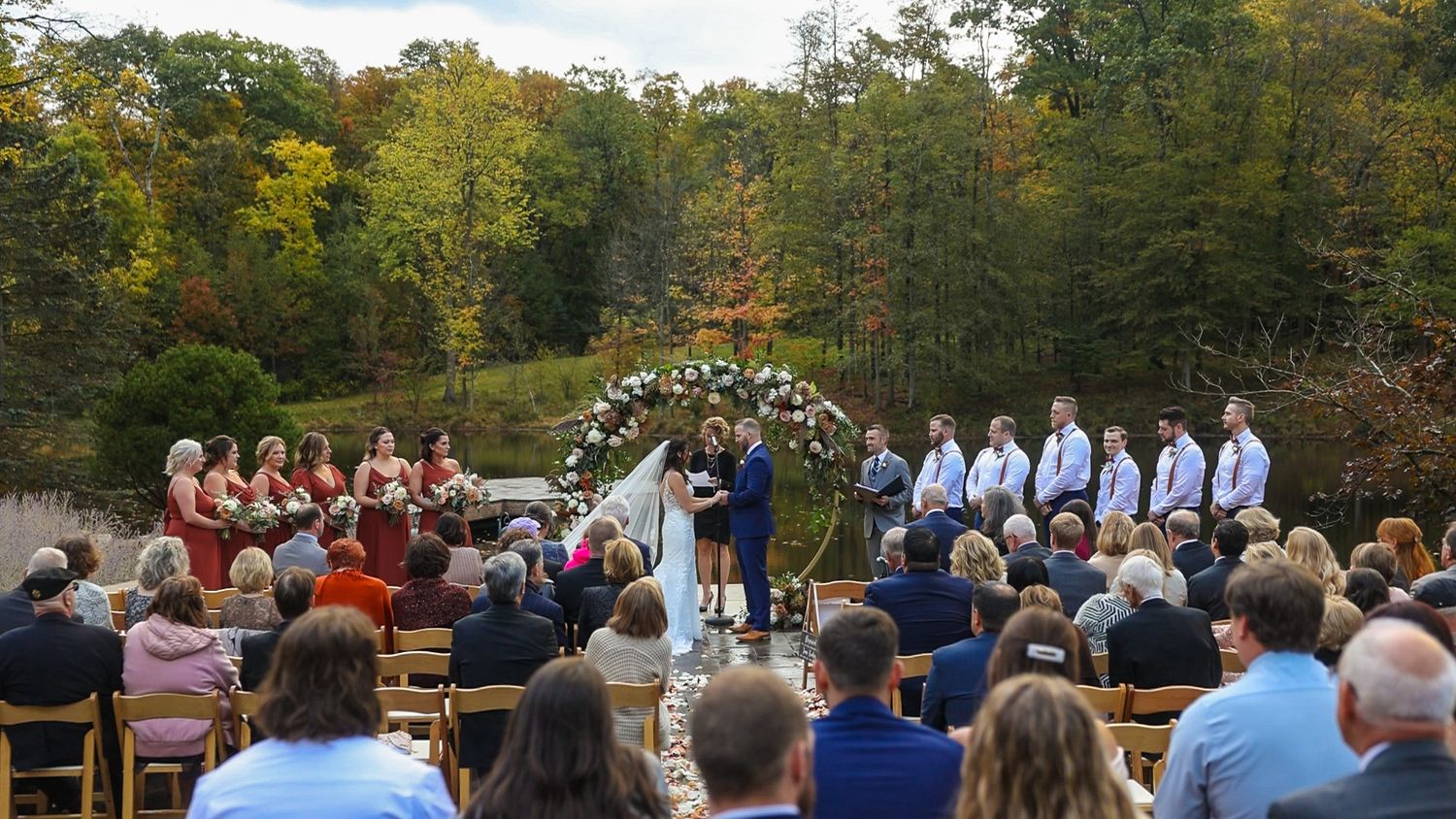Fall outdoor wedding ceremony with guests seated on wooden chairs surrounding a floral arch in a wooded setting.