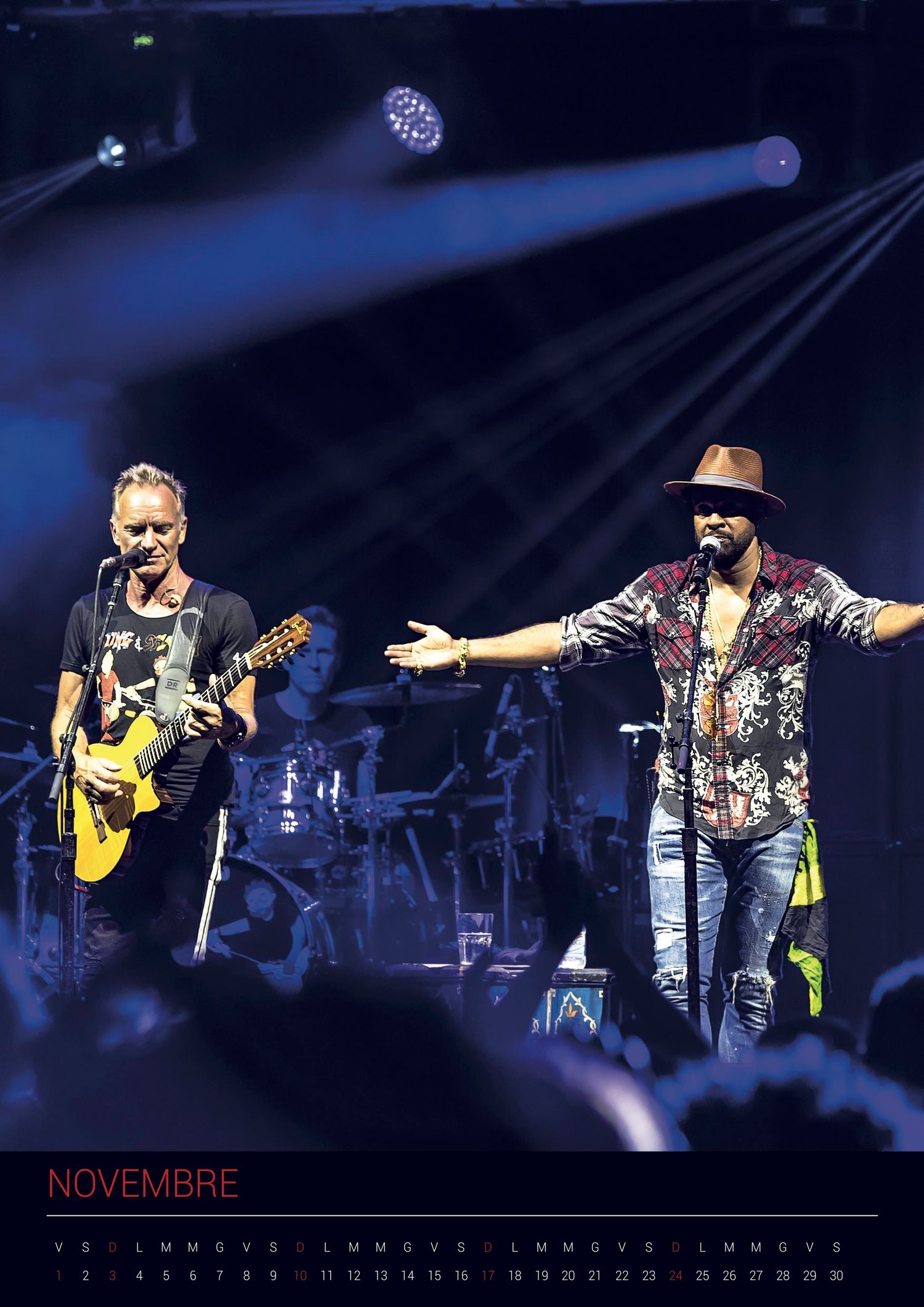 Two performers on stage with one playing guitar and another in floral shirt and hat gesturing during concert with blue lighting.
