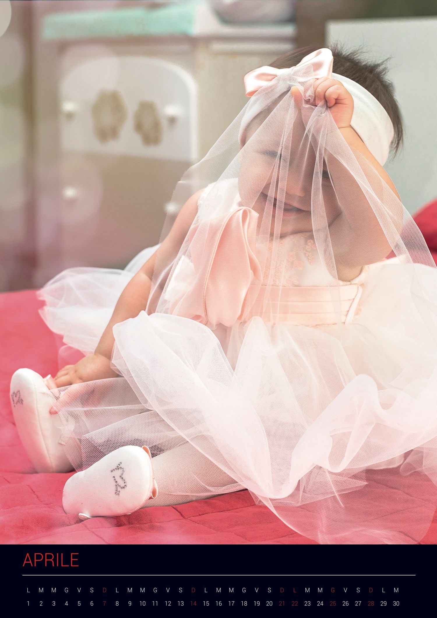 Baby wearing a delicate white tulle veil and pink dress lies on bright red bedding with a calendar showing April.