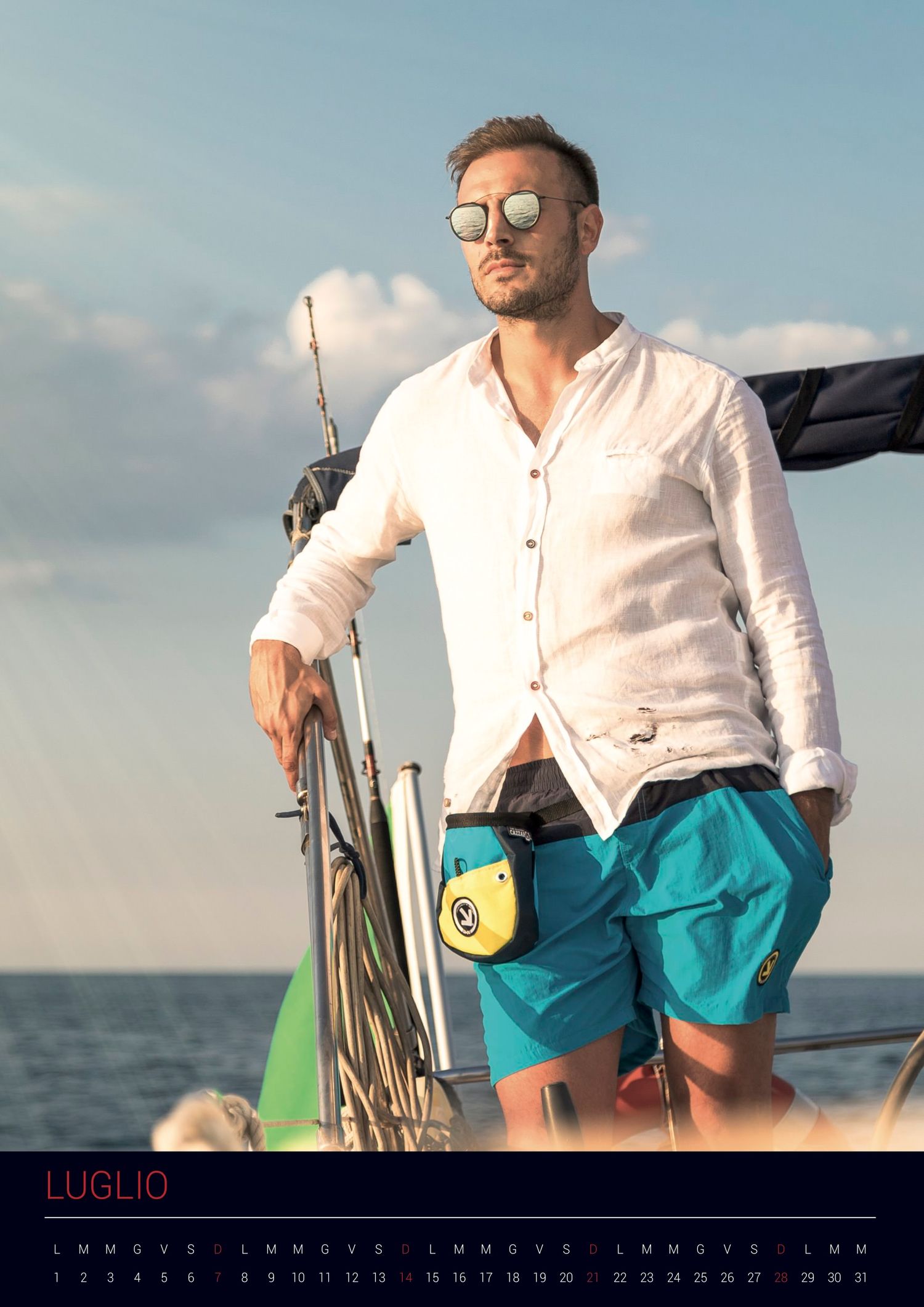 Person in a white shirt and teal shorts stands on a sailboat against a blue sky and ocean horizon.