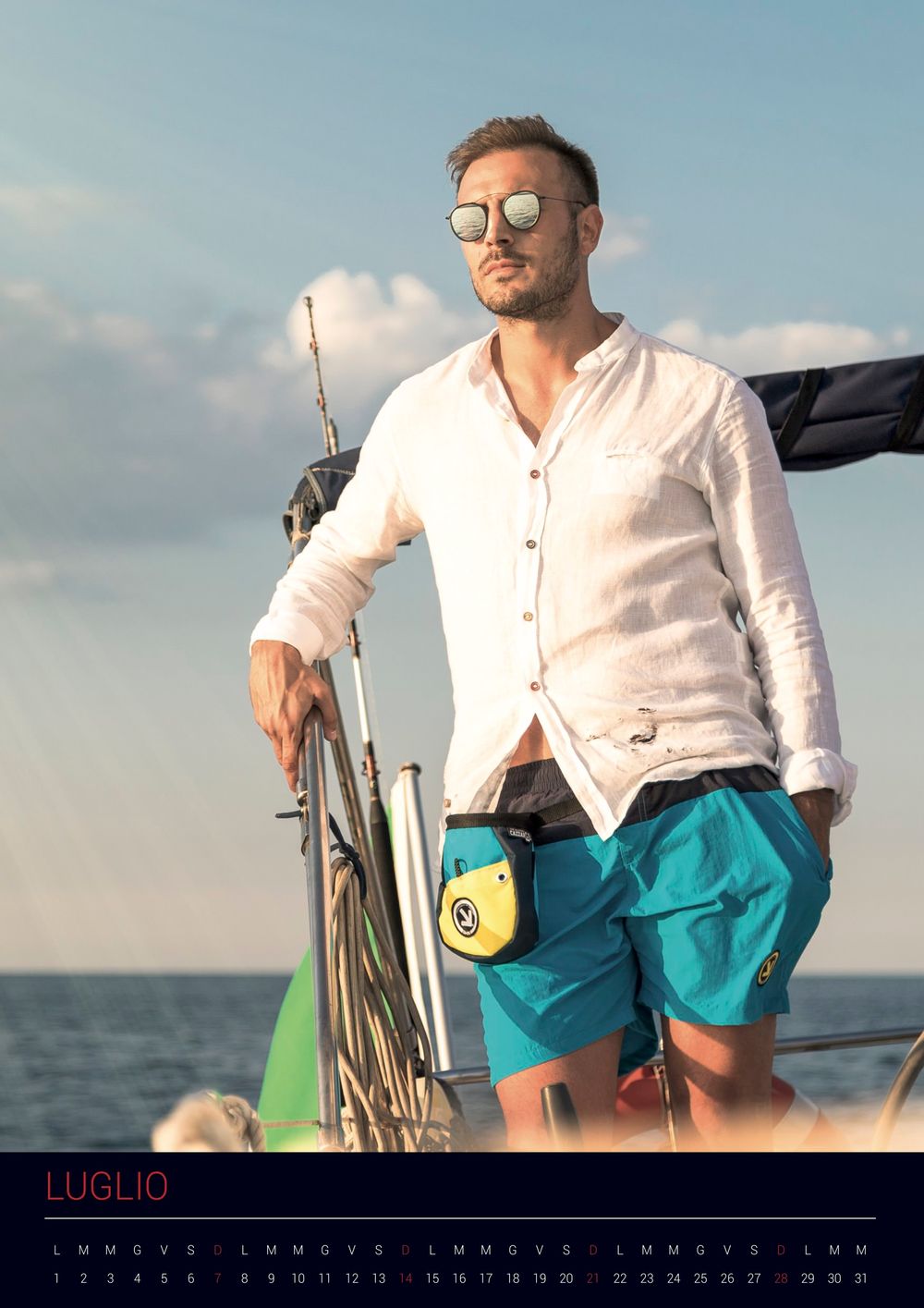 Person in a white shirt and teal shorts stands on a sailboat against a blue sky and ocean horizon.
