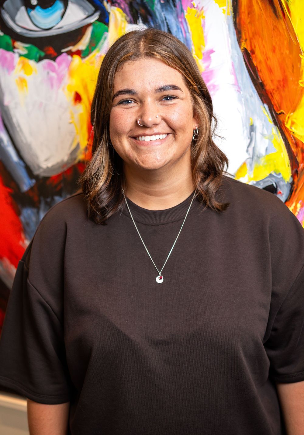 A person wearing a brown shirt smiles while standing in front of a colorful mural with an eye design.