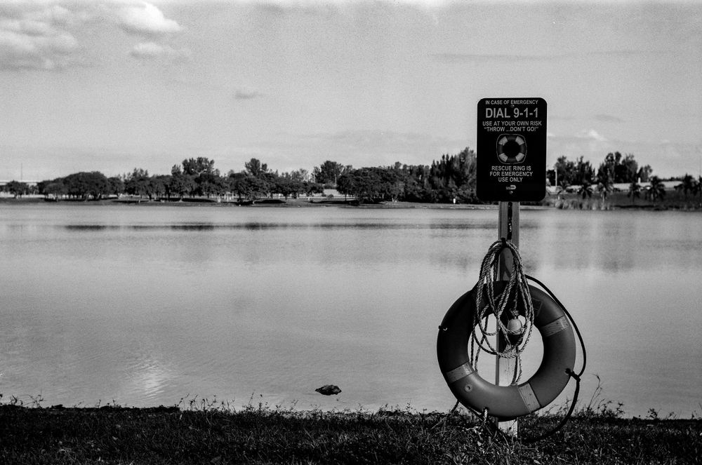 Black and white photo of a life preserver ring and safety sign next to a calm lake.